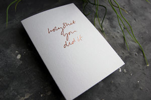 Luxury white greetings card on a grey background. The card has a handwritten phrase hot foiled in a rose gold colour. The card says Holy Shit You Did It..