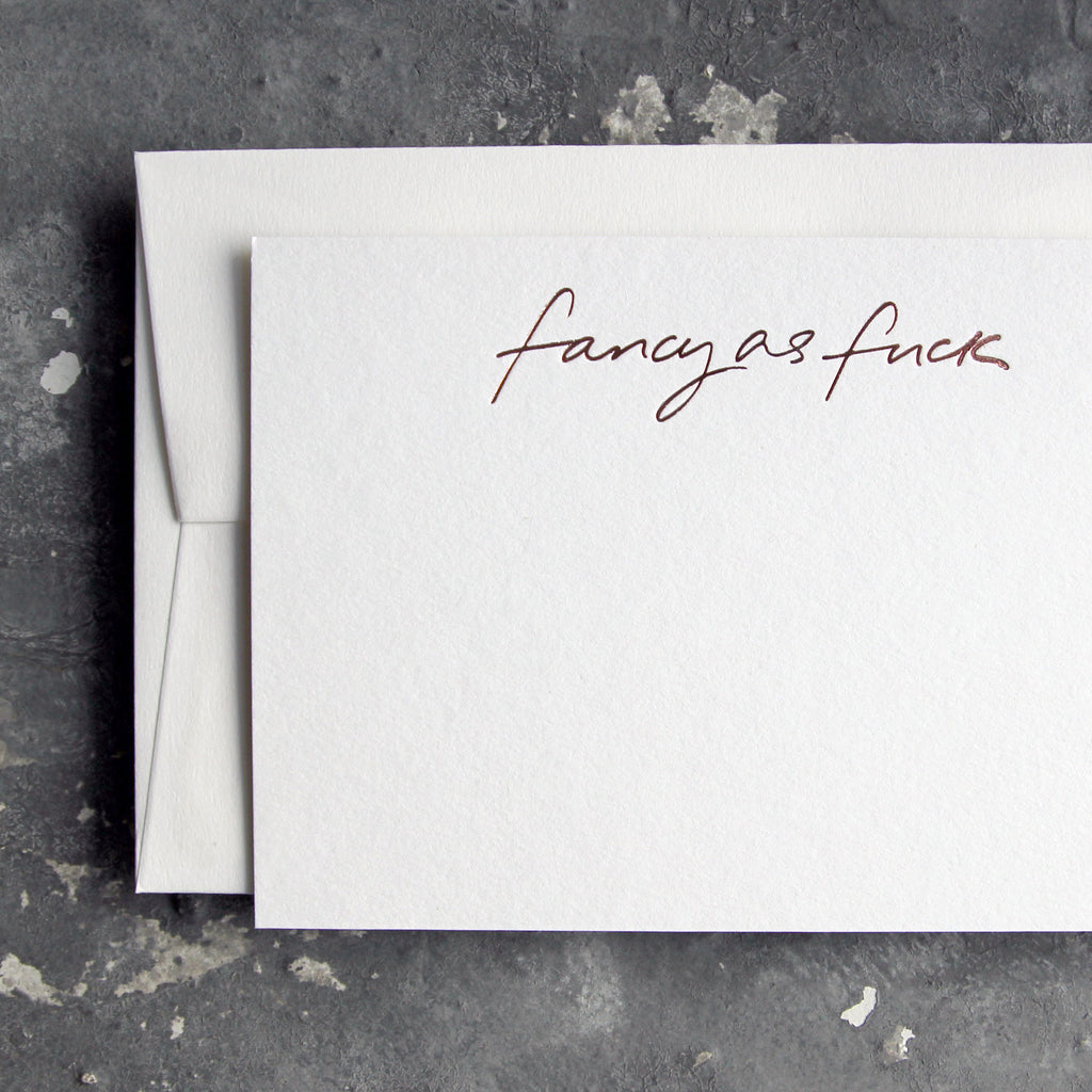 Luxury white notecard and envelope with "Fancy As Fuck" handwritten on the front and hand printed in rose gold foil on a grey background.