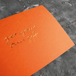 This cash card says Get Your Own Gift and is handwritten and hand printed in gold foil on orange luxury paper on grey board