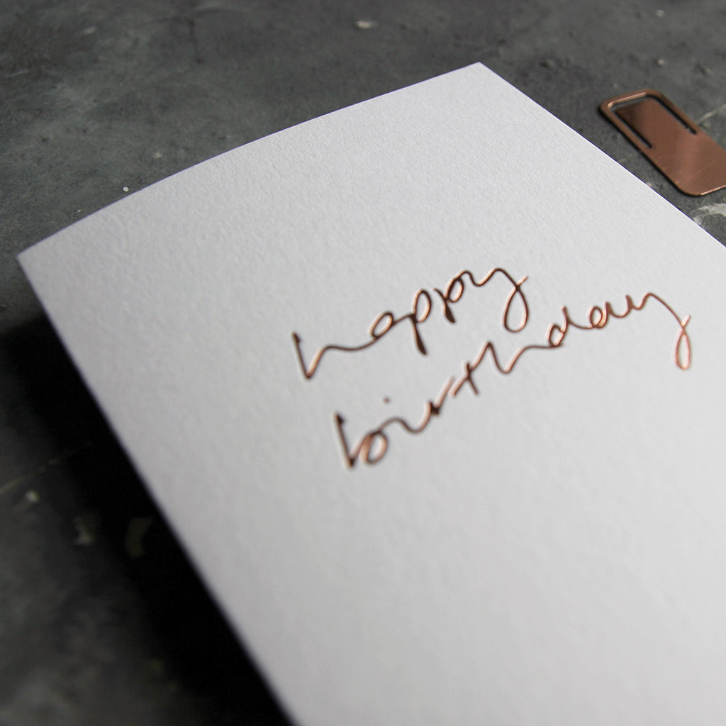 This birthday card says Happy Birthday is handwritten design and handfoiled in rose gold foil on luxury white paper, photographed on a grey board 