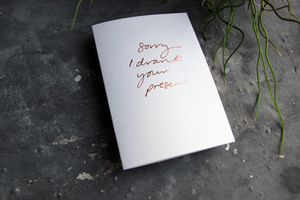 Luxury white greetings card on a grey background. The card has a handwritten phrase hot foiled in a rose gold colour. The card says Sorry I Drank Your Present.