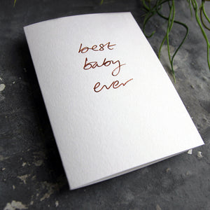 Luxury white greetings card with "Best Baby Ever" handwritten in the front and hand printed in rose gold foil on a grey background with some green plant leaves at the side.