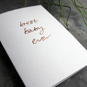 Close up of luxury white greetings card with "Best Baby Ever" handwritten in the front and hand printed in rose gold foil on a grey background with some green plant leaves at the side.