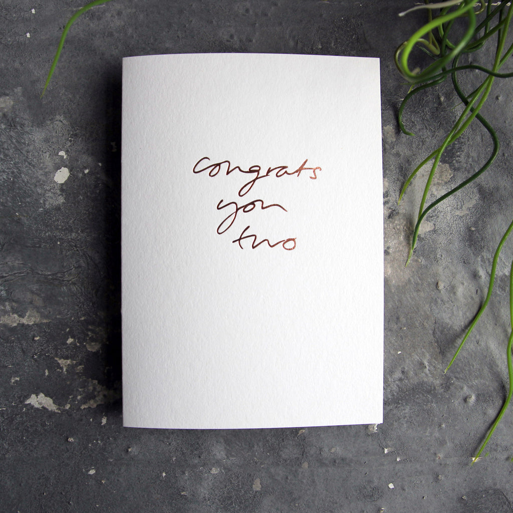 Luxury white greetings card with "Congrats You Two' handwritten in the front and hand printed in rose gold foil on a grey background with some green plant leaves at the side.