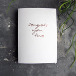Luxury white greetings card with "Congrats You Two' handwritten in the front and hand printed in rose gold foil on a grey background with some green plant leaves at the side.