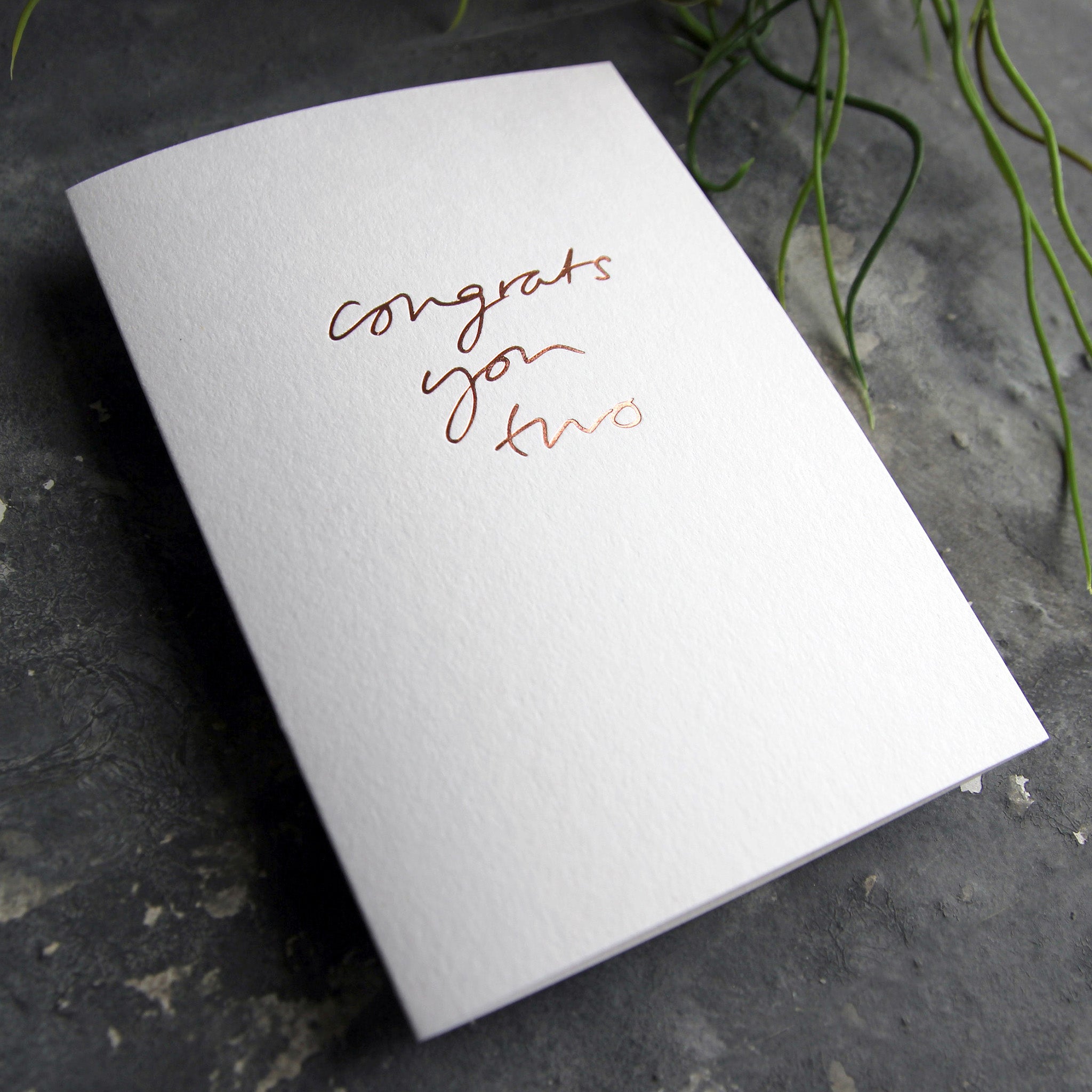 Luxury white greetings card with "Congrats You Two' handwritten in the front and hand printed in rose gold foil on a grey background with some green plant leaves at the side.