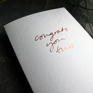 Close up of a luxury white greetings card with "Congrats You Two' handwritten in the front and hand printed in rose gold foil on a grey background with some green plant leaves at the side.