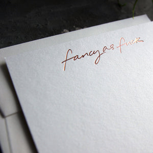 Close up of luxury white notecard and envelope with "Fancy As Fuck" handwritten on the front and hand printed in rose gold foil on a grey background.