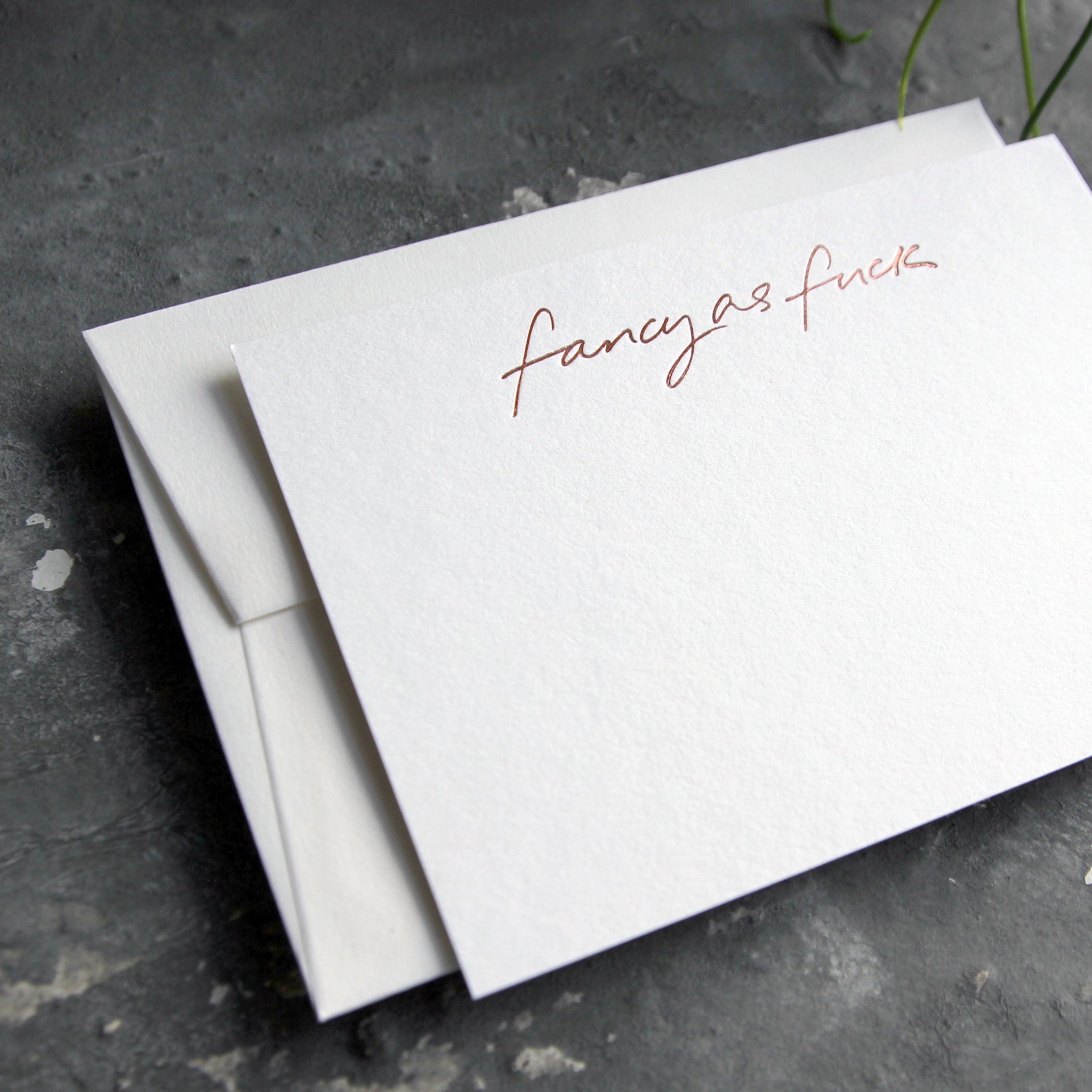 Luxury white notecard and envelope with "Fancy As Fuck" handwritten on the front and hand printed in rose gold foil on a grey background.