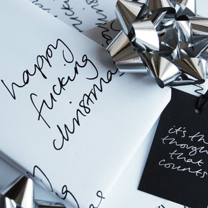 This luxury wrapping paper has 'Happy Fucking Christmas' handwritten and printed in black and white. The wrapped gift has a hand foiled gift tag which says It's The Thought That Counts. Photographed next to silver bows.