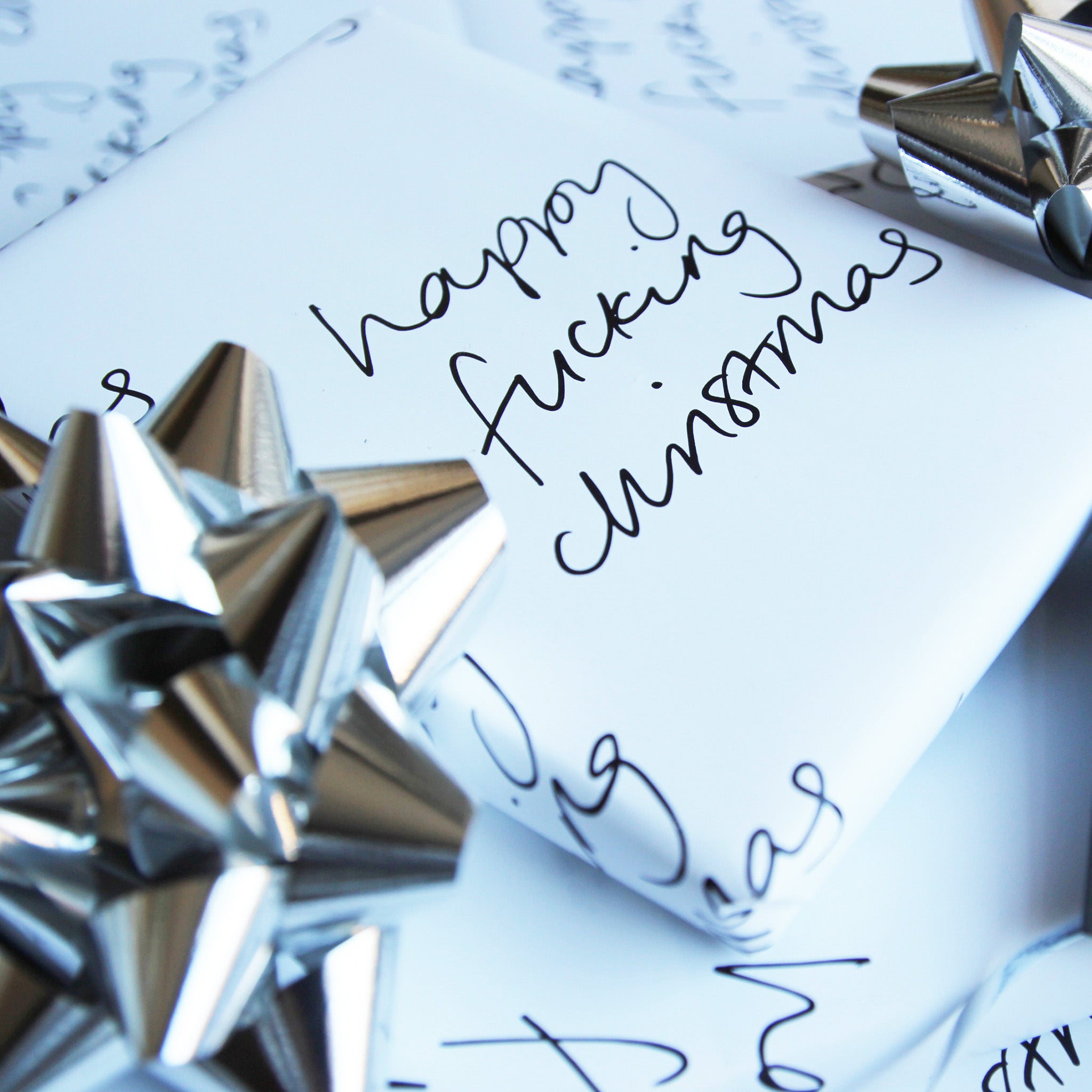 This luxury wrapping paper has 'Happy Fucking Christmas' handwritten and printed in black and white. The wrapped gift has a hand foiled gift tag which says It's The Thought That Counts. Photographed next to silver bows.