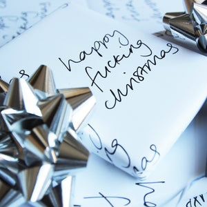 This luxury wrapping paper has 'Happy Fucking Christmas' handwritten and printed in black and white. The wrapped gift has a hand foiled gift tag which says It's The Thought That Counts. Photographed next to silver bows.