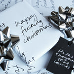 This luxury wrapping paper has 'Happy Fucking Christmas' handwritten and printed in black and white. The wrapped gift has a hand foiled gift tag which says It's The Thought That Counts. Photographed next to silver bows.