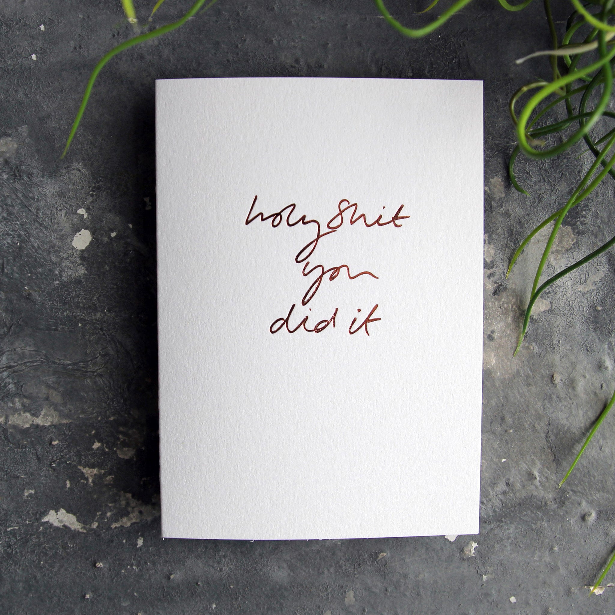 Luxury white greetings card with "Holy Shit You Did It' handwritten in the front and hand printed in rose gold foil on a grey background with some green plant leaves at the side.