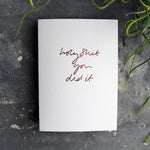 Luxury white greetings card with "Holy Shit You Did It' handwritten in the front and hand printed in rose gold foil on a grey background with some green plant leaves at the side.