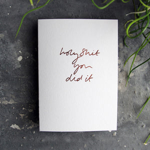 Luxury white greetings card with "Holy Shit You Did It' handwritten in the front and hand printed in rose gold foil on a grey background with some green plant leaves at the side.