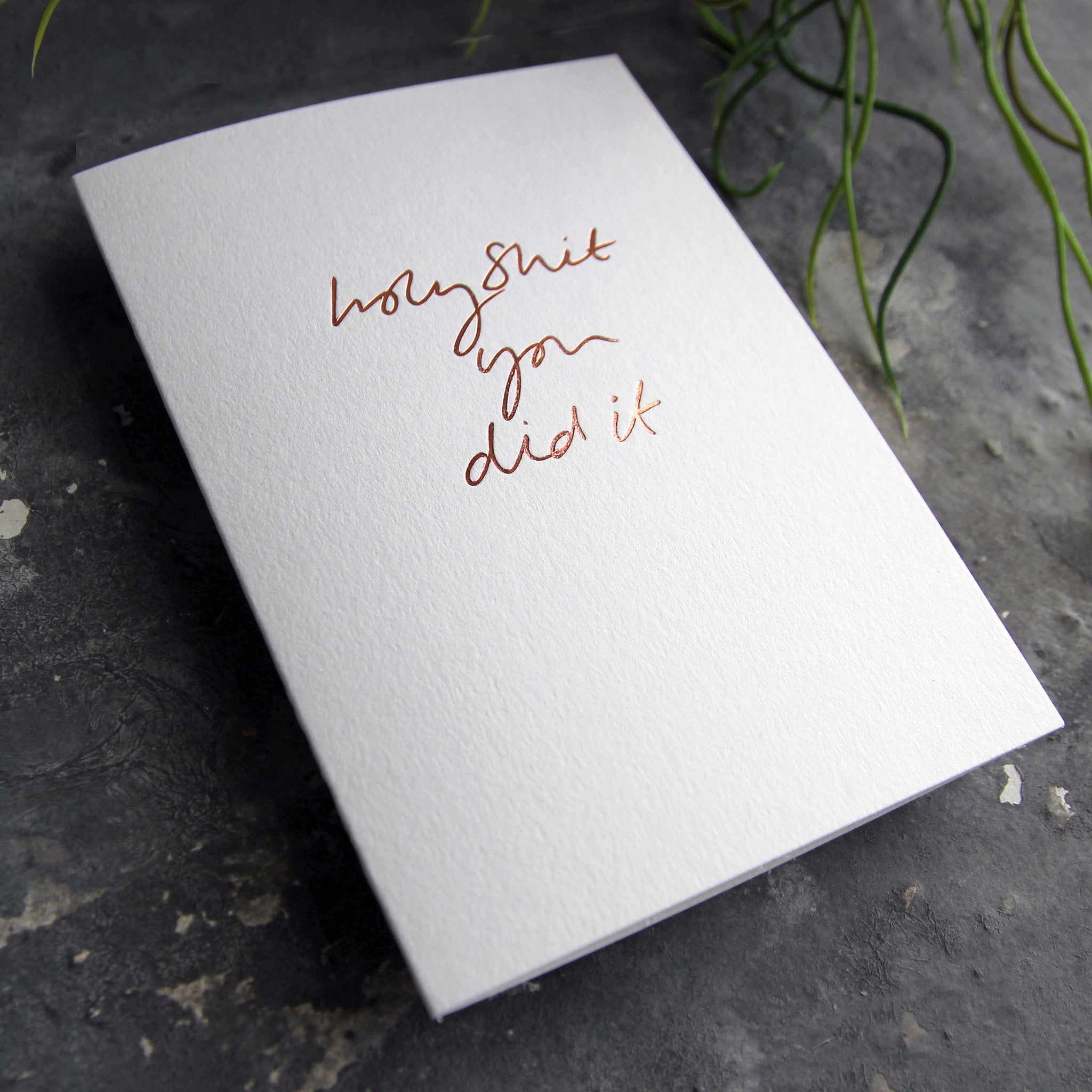 Luxury white greetings card with "Holy Shit You Did It' handwritten in the front and hand printed in rose gold foil on a grey background with some green plant leaves at the side.