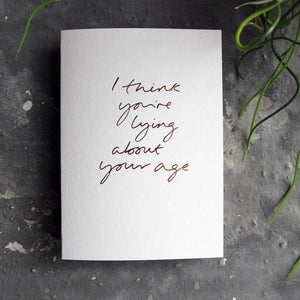 Luxury white greetings card with "I Think You're Lying About Your Age" handwritten in the front and hand printed in rose gold foil on a grey background with some green plant leaves at the side.