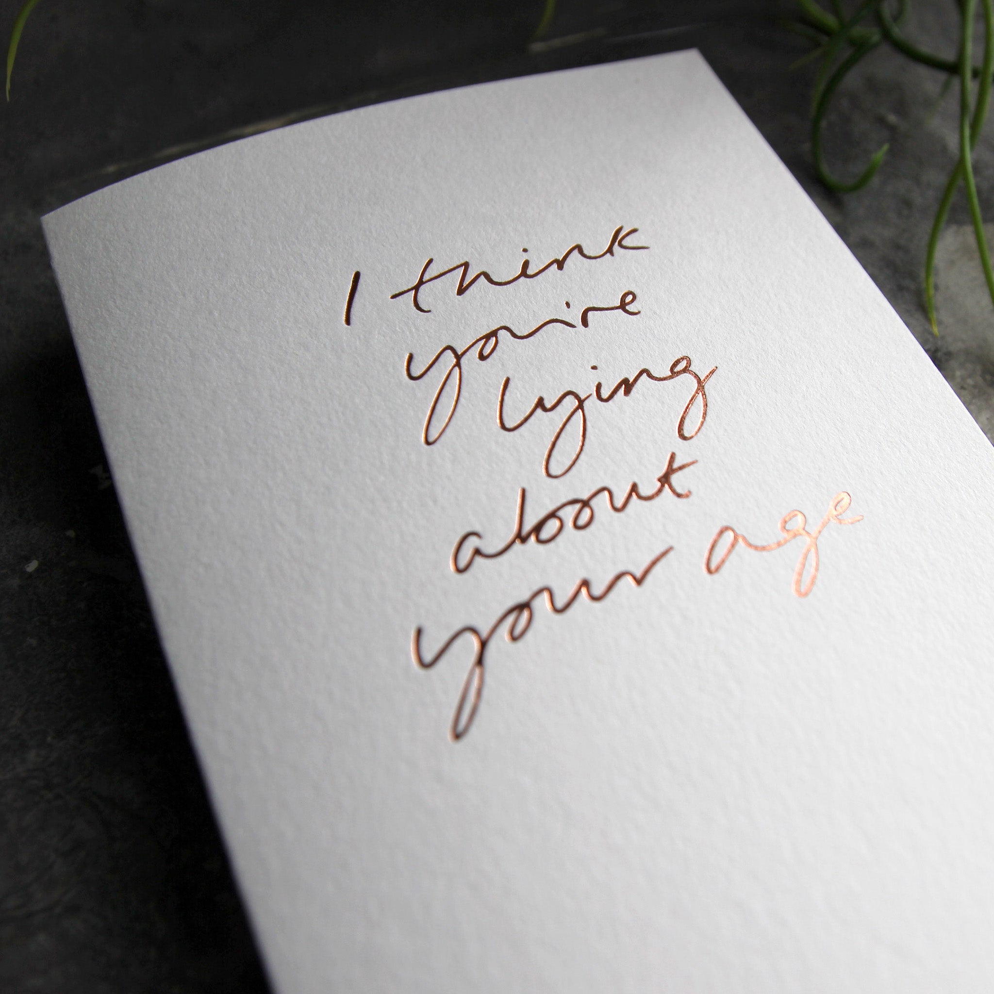 Close up of luxury white greetings card with "I Think You're Lying About Your Age" handwritten in the front and hand printed in rose gold foil on a grey background with some green plant leaves at the side.