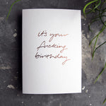 Luxury white greetings card with "It's Your Fucking Birthday" handwritten in the front and hand printed in rose gold foil on a grey background with some green plant leaves at the side.