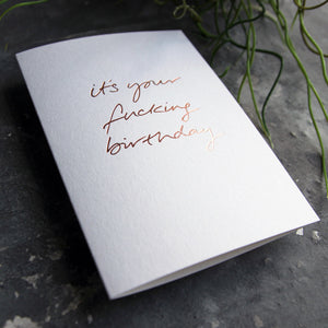 Luxury white greetings card with "It's Your Fucking Birthday" handwritten in the front and hand printed in rose gold foil on a grey background with some green plant leaves at the side.