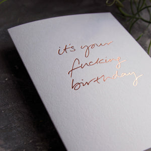 Close up of a luxury white greetings card with "It's Your Fucking Birthday" handwritten in the front and hand printed in rose gold foil on a grey background with some green plant leaves at the side.
