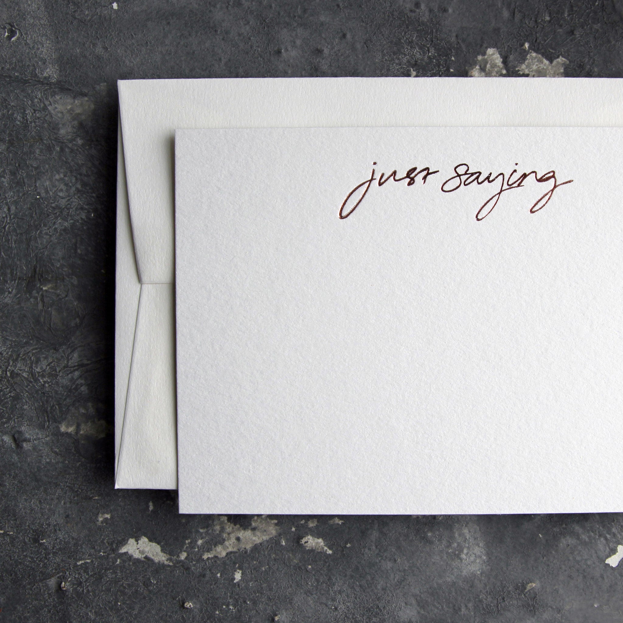 Luxury white notecard and envelope with "Just Saying" handwritten on the front and hand printed in rose gold foil on a grey background.