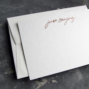 Luxury white notecard and envelope with "Just Saying" handwritten on the front and hand printed in rose gold foil on a grey background.