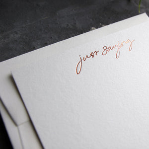 Close up of a luxury white notecard and envelope with "Just Saying" handwritten on the front and hand printed in rose gold foil on a grey background.