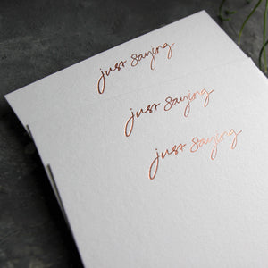 Close up of 3 luxury white notecards with "Just Saying" handwritten on the front and hand printed in rose gold foil on a grey background.