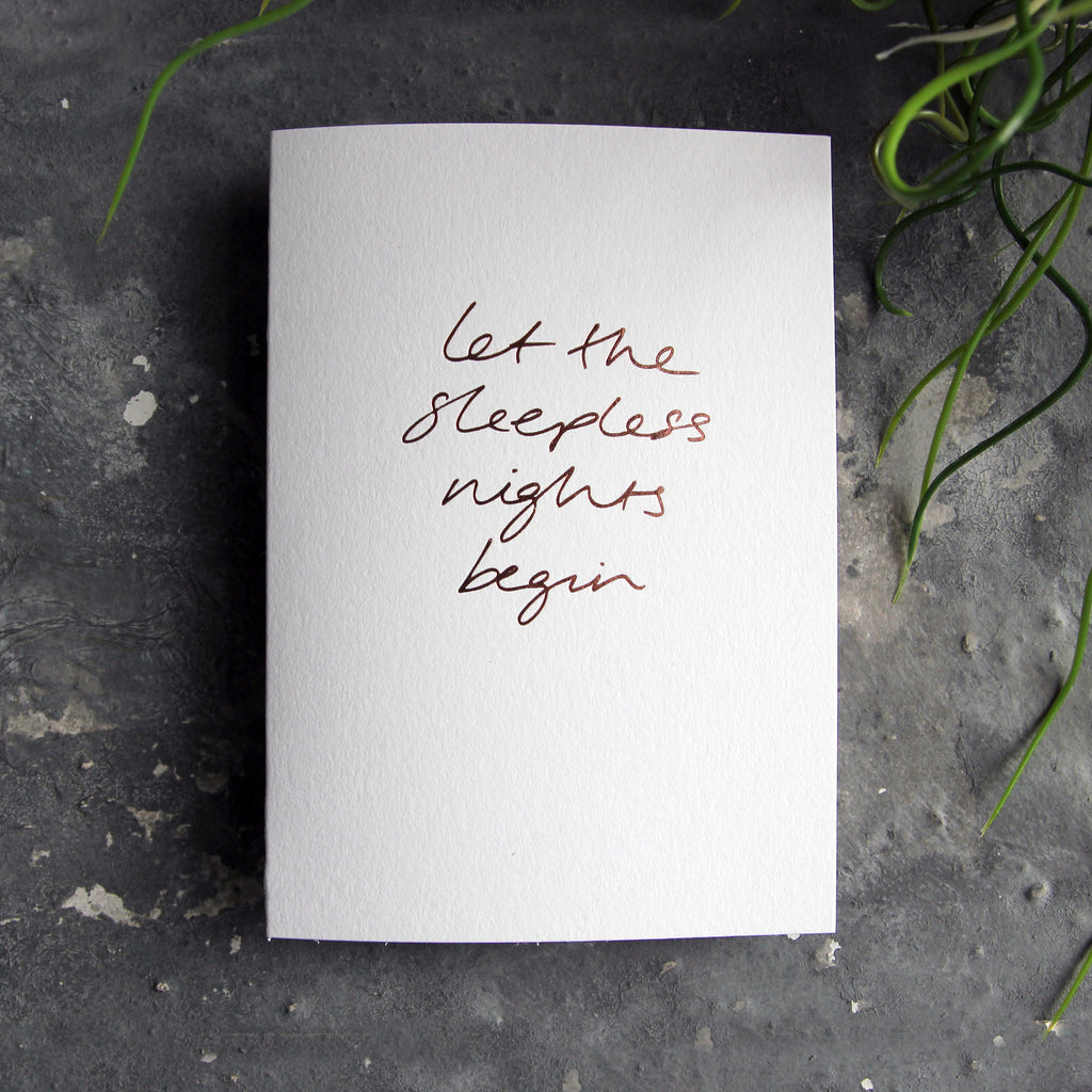 Luxury white greetings card with "Let The Sleepless Nights Begin" handwritten in the front and hand printed in rose gold foil on a grey background with some green plant leaves at the side.