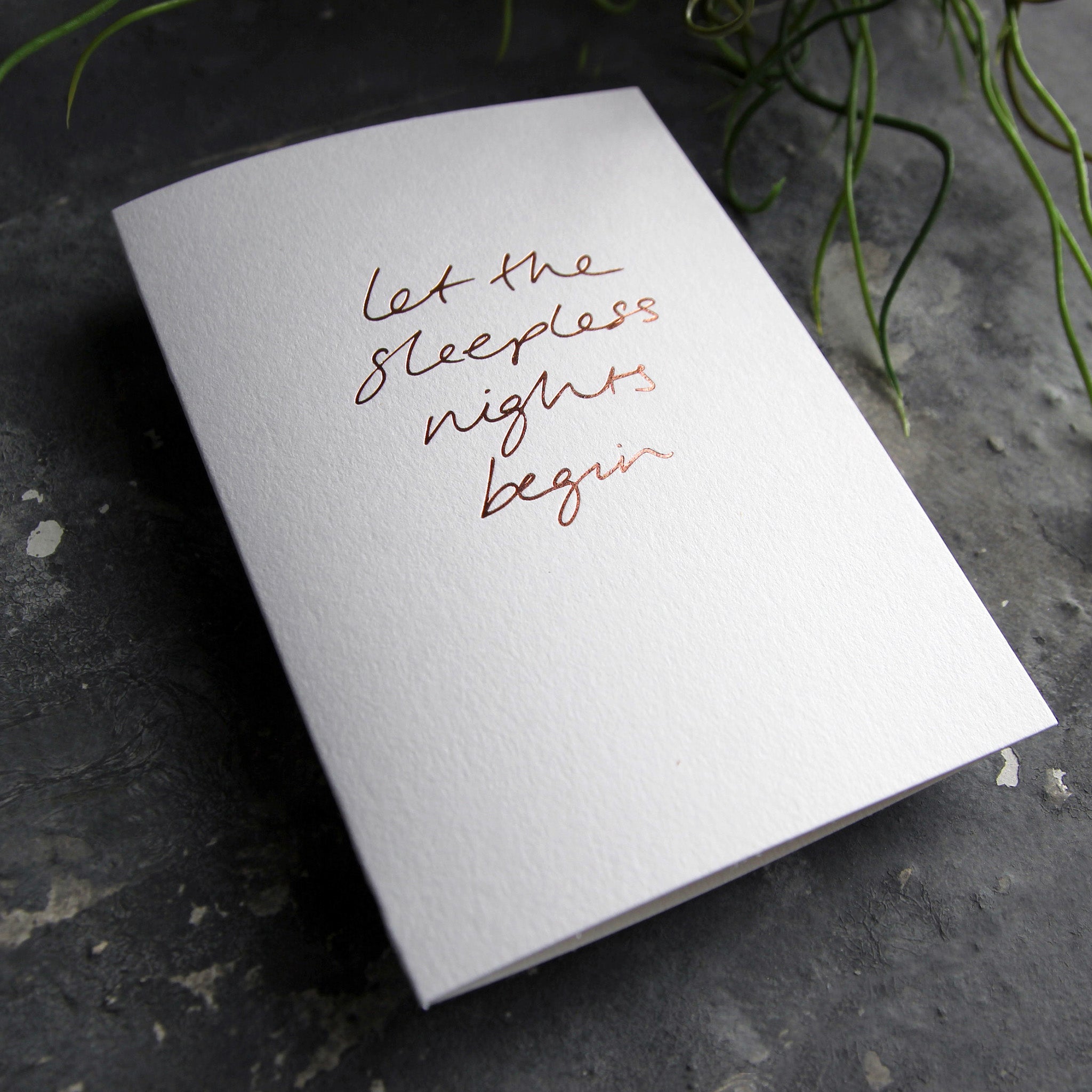 Luxury white greetings card with "Let The Sleepless Nights Begin" handwritten in the front and hand printed in rose gold foil on a grey background with some green plant leaves at the side.