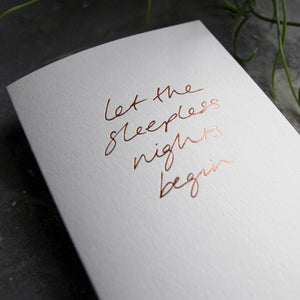 Close up of luxury white greetings card with "Let The Sleepless Nights Begin" handwritten in the front and hand printed in rose gold foil on a grey background with some green plant leaves at the side.