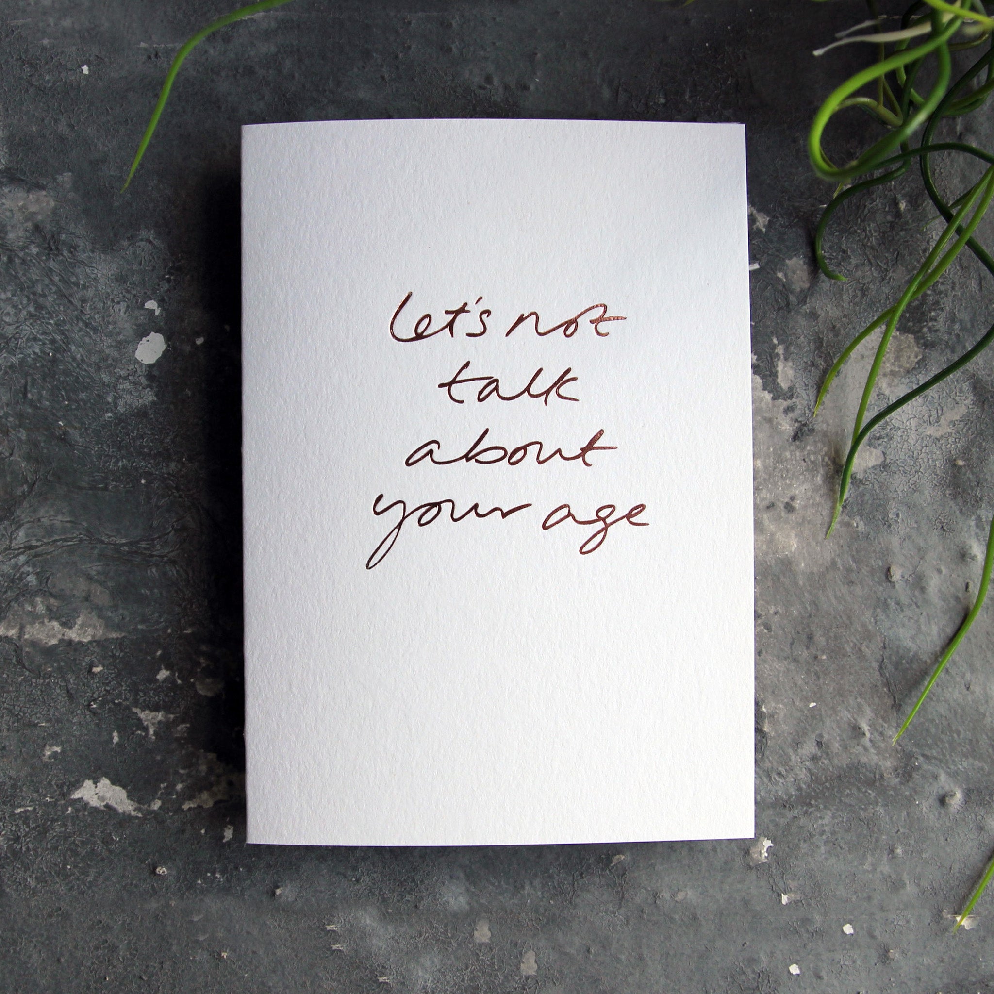 Luxury white greetings card with "Let's Not Talk About Your Age' handwritten in the front and hand printed in rose gold foil on a grey background with some green plant leaves at the side.