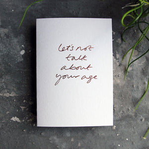 Luxury white greetings card with "Let's Not Talk About Your Age' handwritten in the front and hand printed in rose gold foil on a grey background with some green plant leaves at the side.