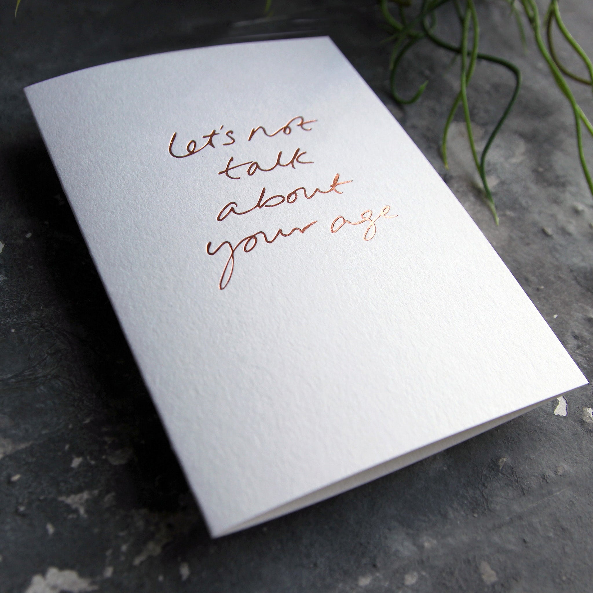 Luxury white greetings card with "Let's Not Talk About Your Age' handwritten in the front and hand printed in rose gold foil on a grey background with some green plant leaves at the side.