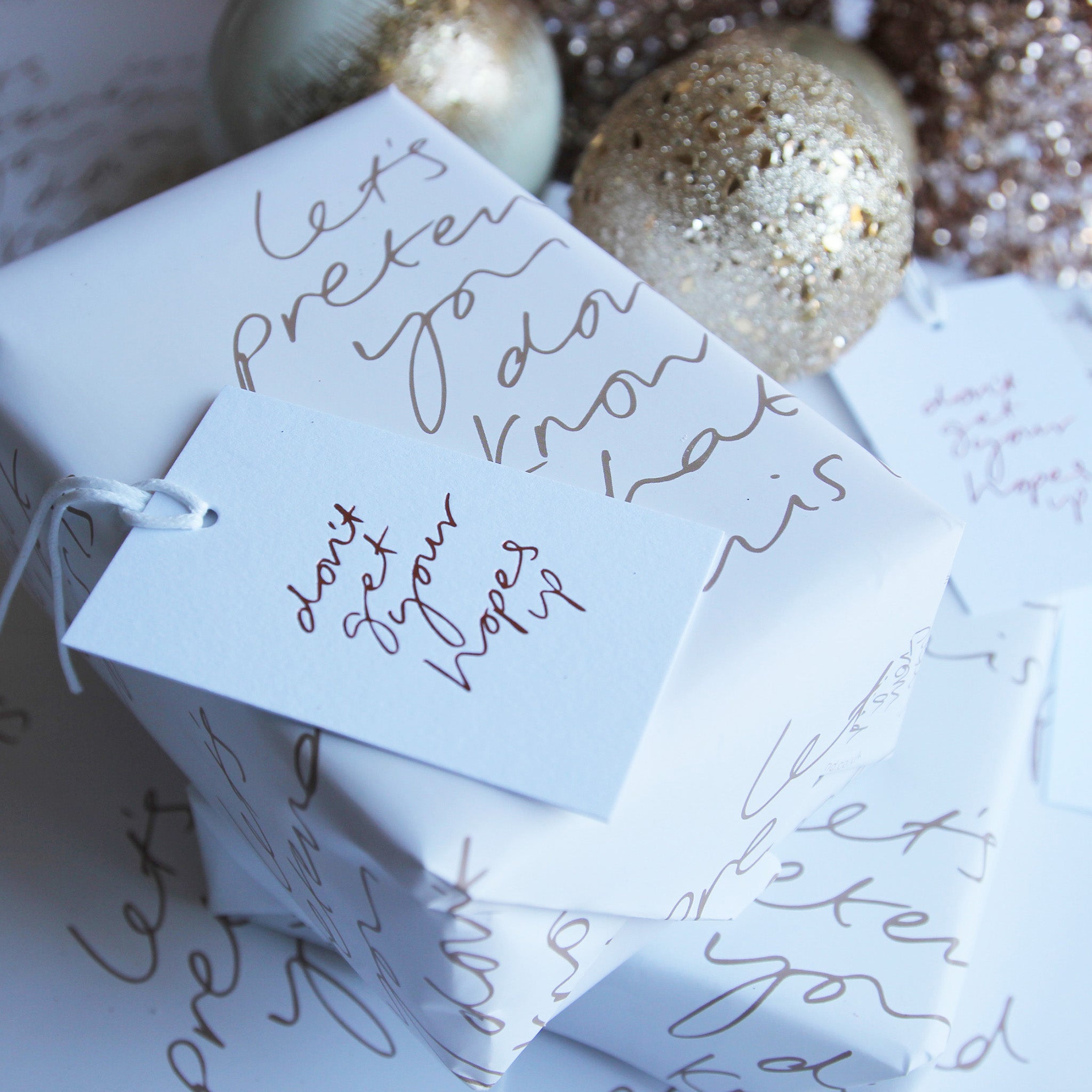 This luxury wrapping paper has "Let's pretend you don;t know what this is" handwritten and printed in a bronze colour. The wrapped gift has a hand foiled gift tag which says Don;t Get Your Hope Up. Photographed next to gold baubles.