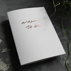 Luxury white greetings card with "Meant To Be" handwritten in the front and hand printed in rose gold foil on a grey background with some green plant leaves at the side.