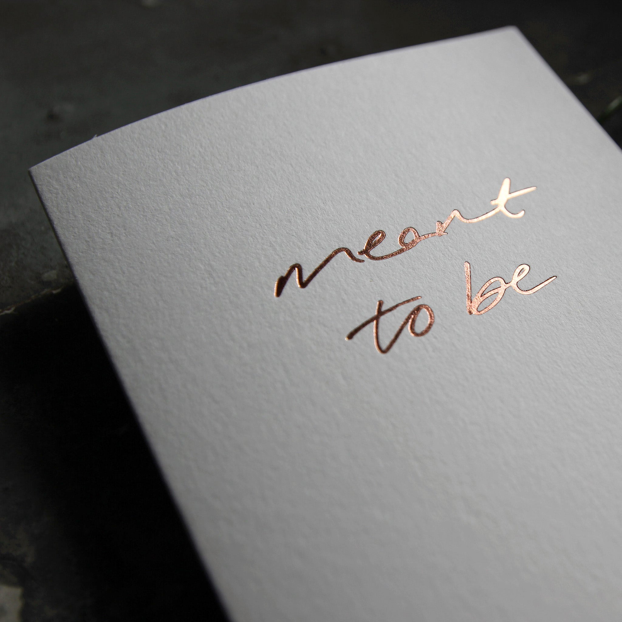 Luxury white greetings card with "Meant To Be" handwritten in the front and hand printed in rose gold foil on a grey background with some green plant leaves at the side.