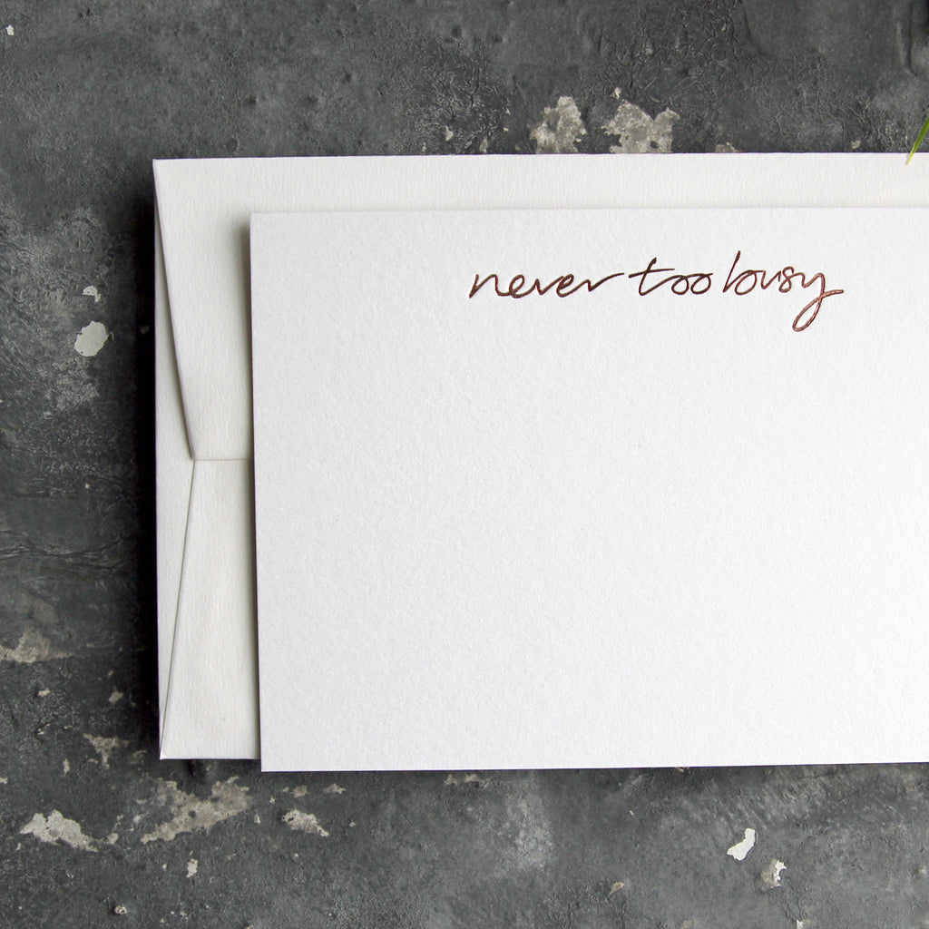 Luxury white notecard and envelope with "Never Too Busy" handwritten on the front and hand printed in rose gold foil on a grey background.