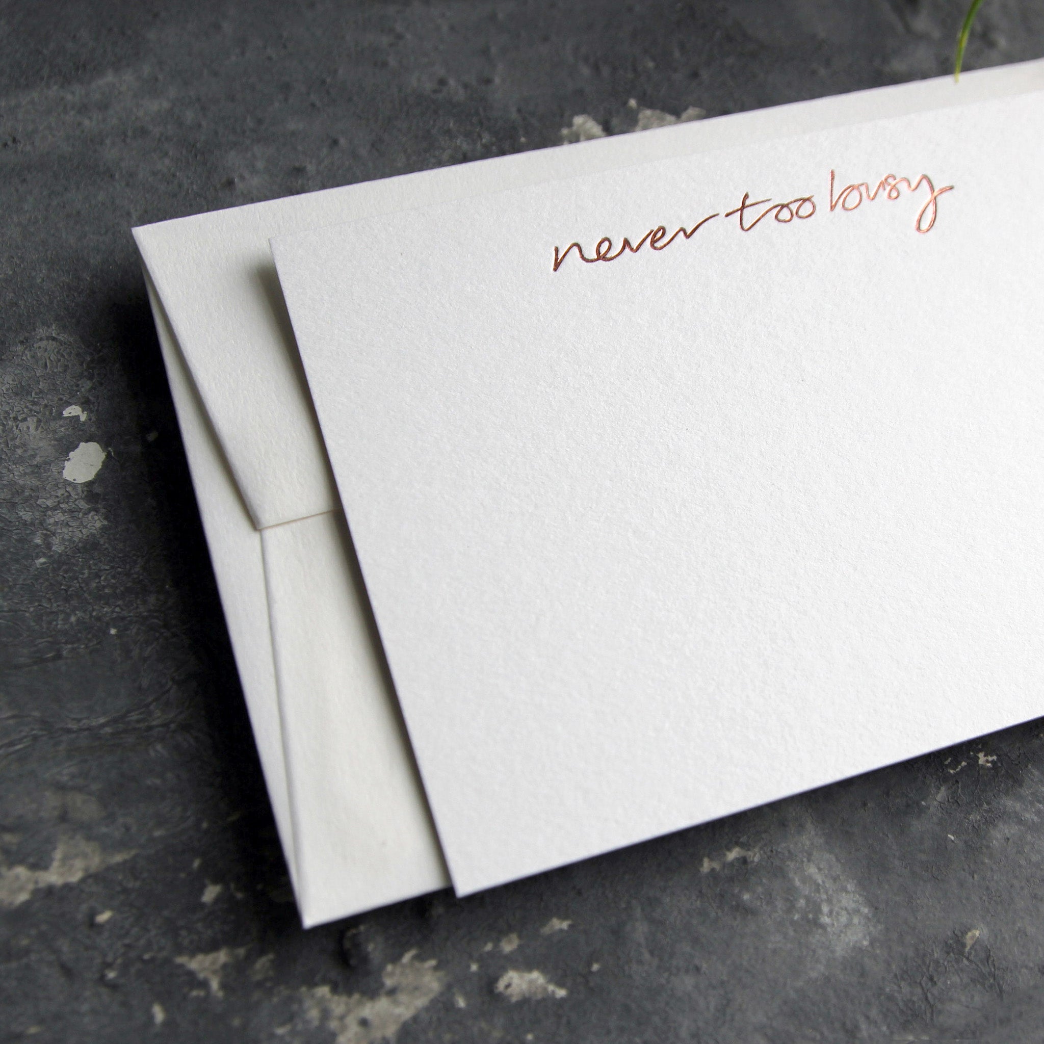 Luxury white notecard and envelope with "Never Too Busy" handwritten on the front and hand printed in rose gold foil on a grey background.