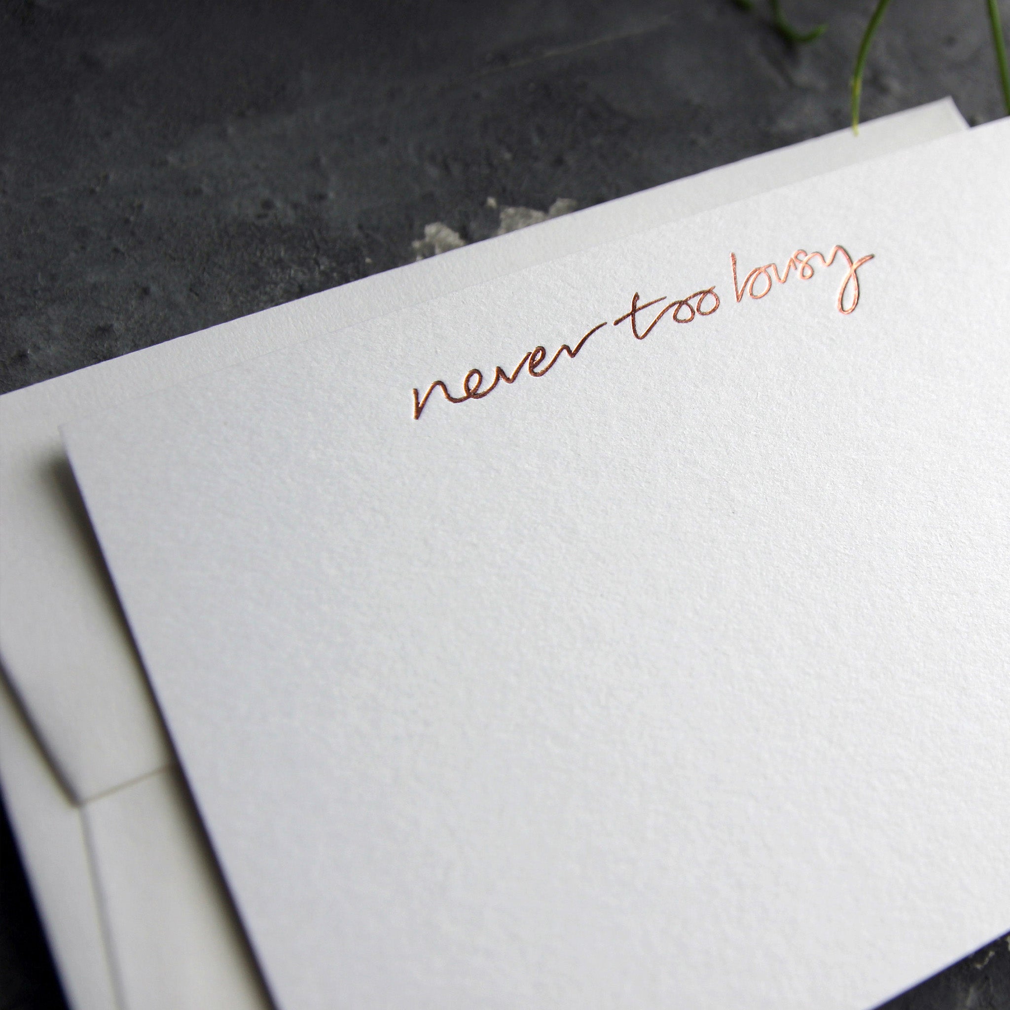 Close up of luxury white notecard and envelope with "Never Too Busy" handwritten on the front and hand printed in rose gold foil on a grey background.