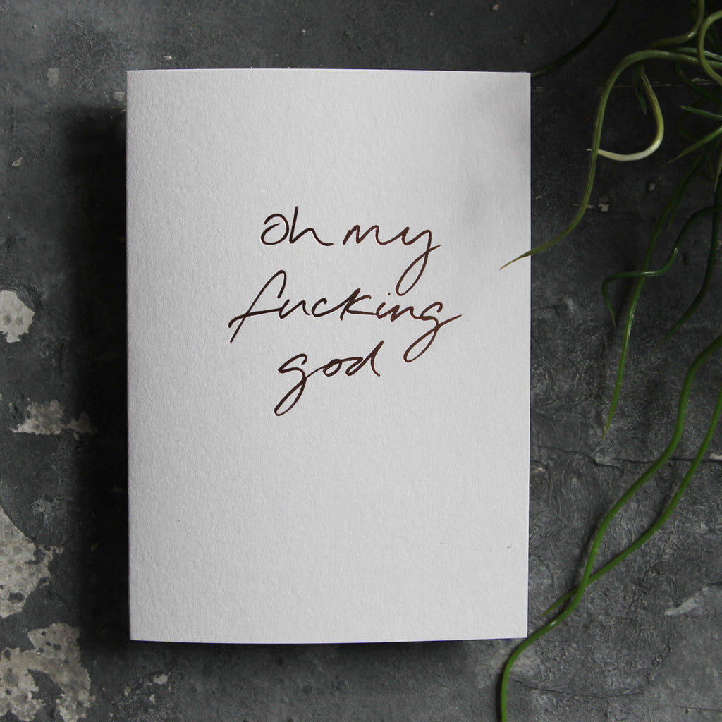 Luxury white greetings card with "Oh My Fucking God" handwritten in the front and hand printed in rose gold foil on a grey background with some green plant leaves at the side.