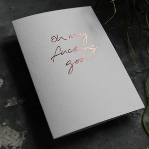 Luxury white greetings card with "Oh My Fucking God" handwritten in the front and hand printed in rose gold foil on a grey background with some green plant leaves at the side.