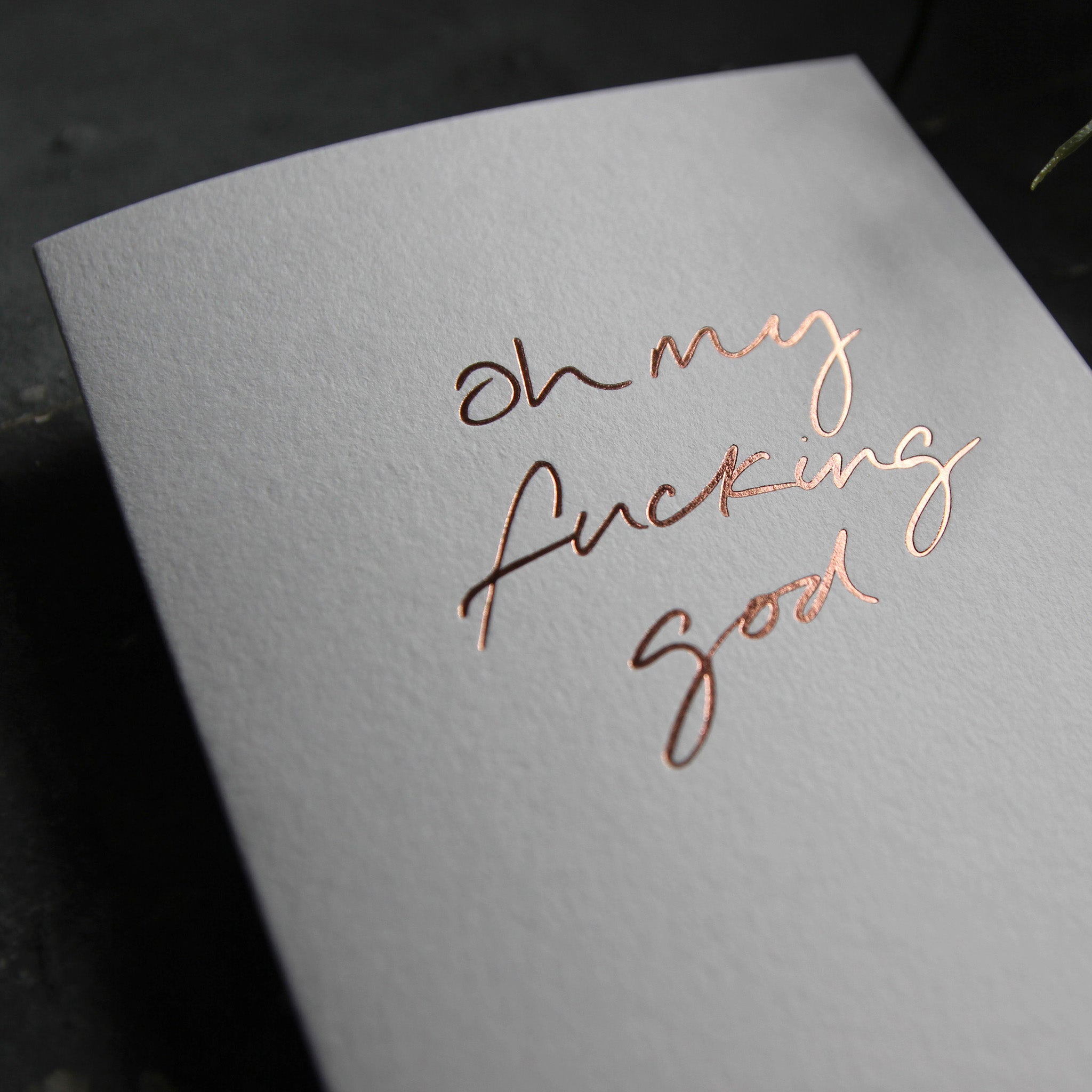 Luxury white greetings card with "Oh My Fucking God" handwritten in the front and hand printed in rose gold foil on a grey background with some green plant leaves at the side.