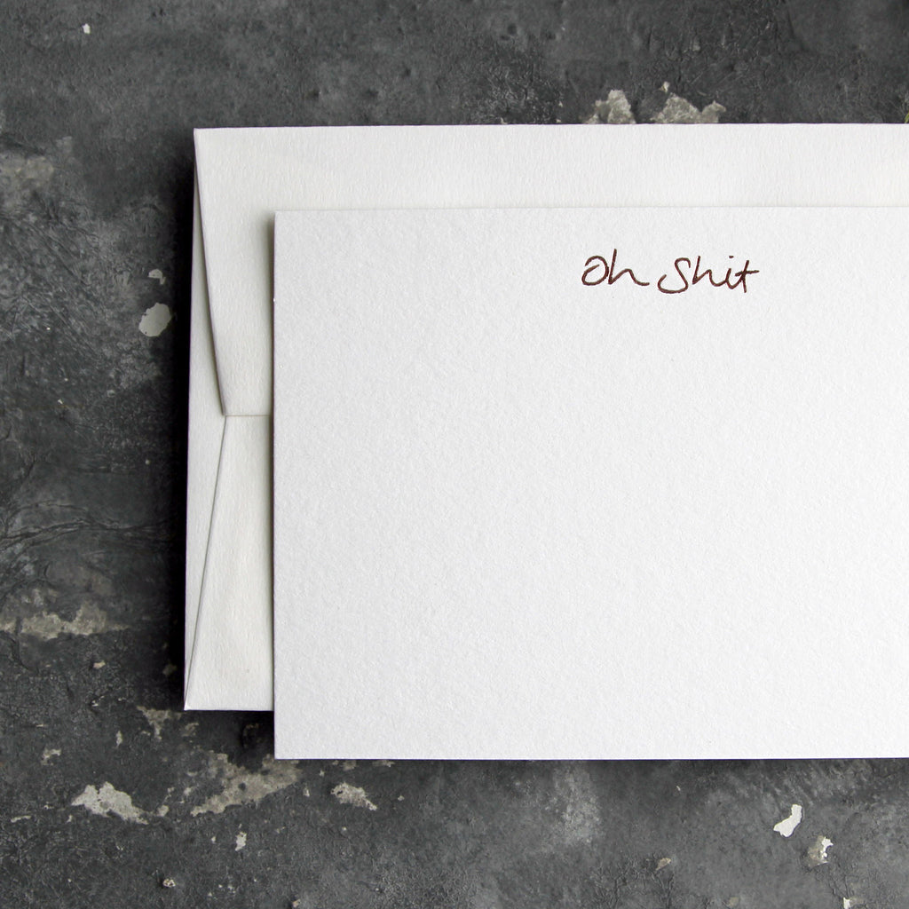 Luxury white notecard and envelope with "Oh Shit" handwritten on the front and hand printed in rose gold foil on a grey background.