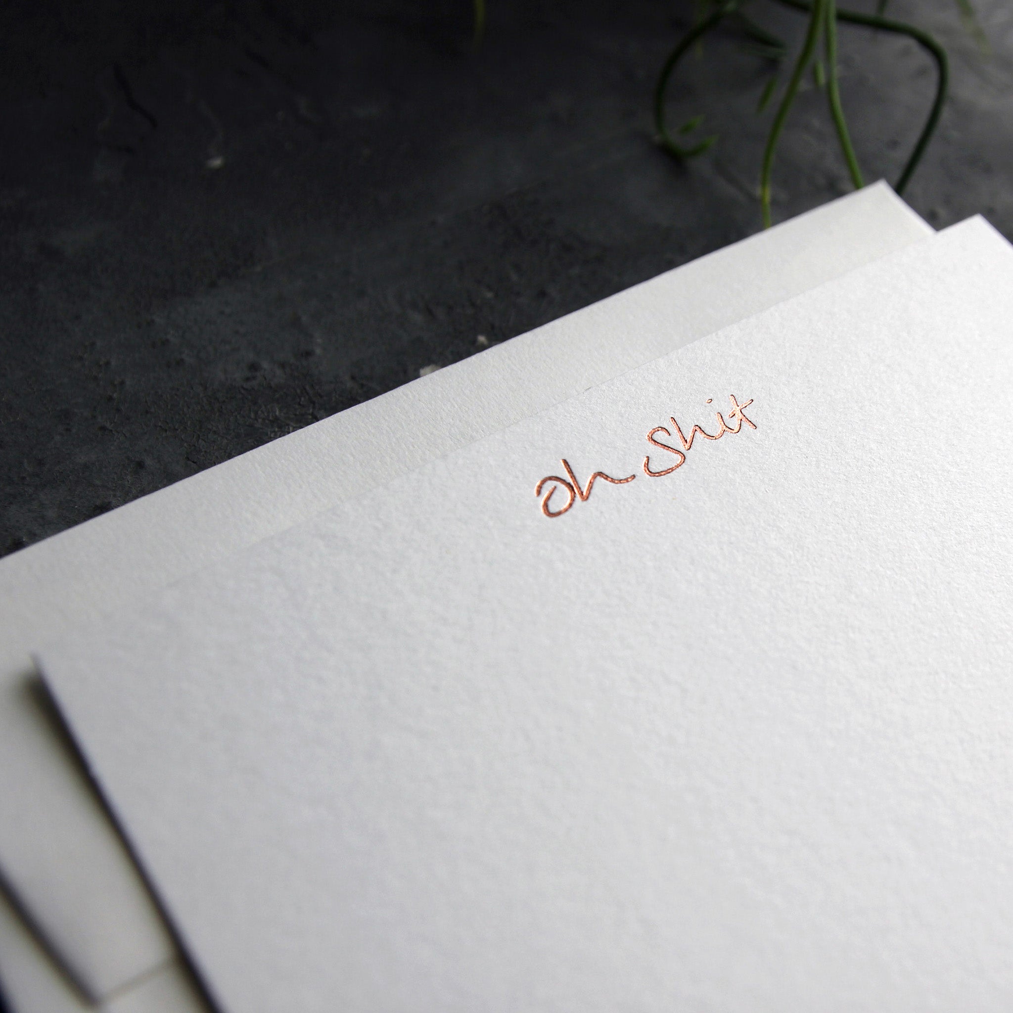 Close up of a luxury white notecard and envelope with "Oh Shit" handwritten on the front and hand printed in rose gold foil on a grey background.