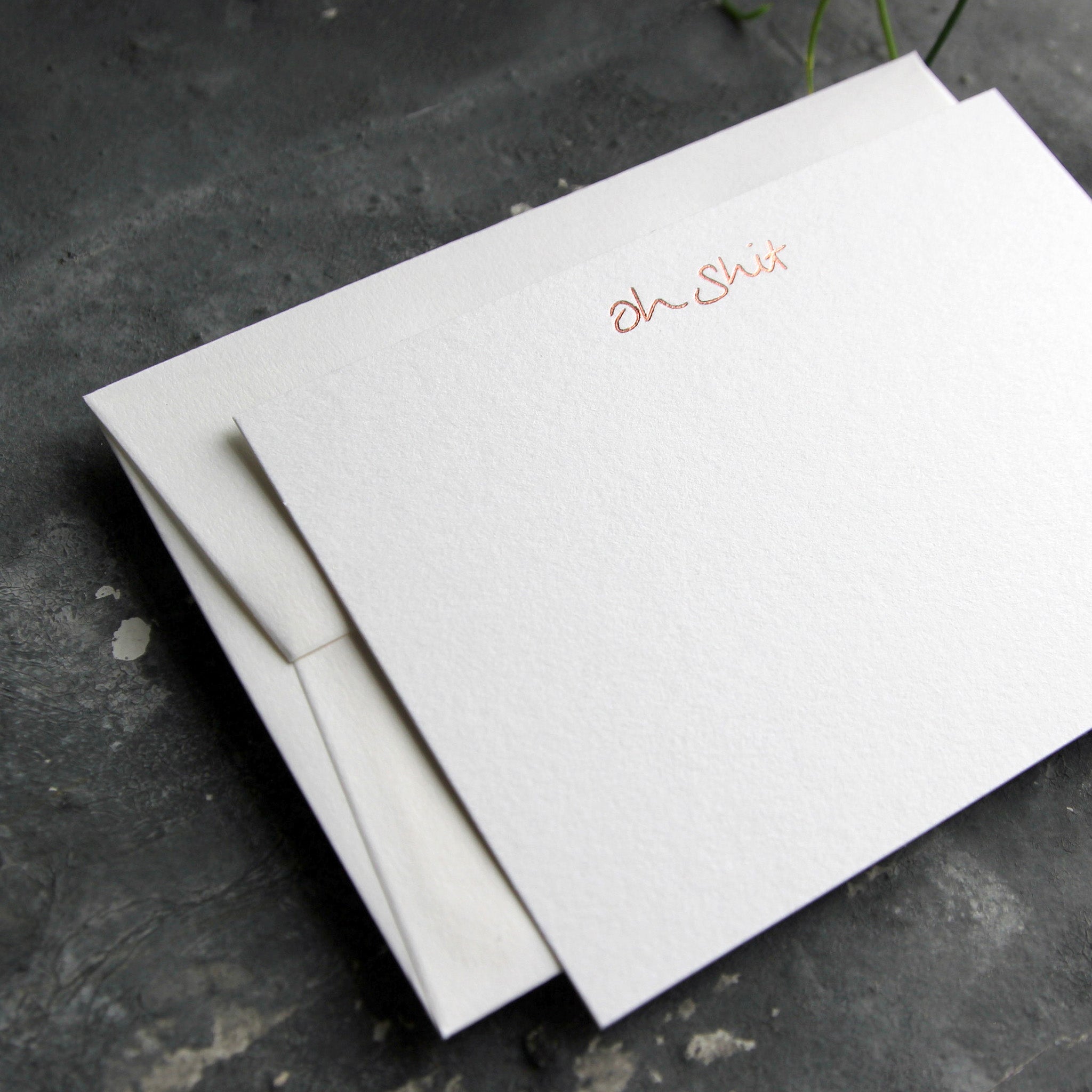 Luxury white notecard and envelope with "Oh Shit" handwritten on the front and hand printed in rose gold foil on a grey background.