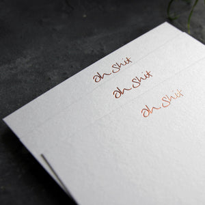 Close up of 3 luxury white notecards with "Oh Shit" handwritten on the front and hand printed in rose gold foil on a grey background.