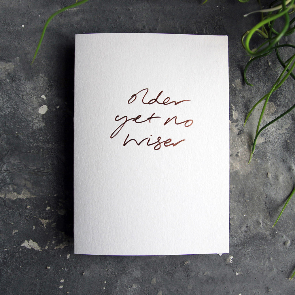 Luxury white greetings card with "Older Yet No Wiser" handwritten in the front and hand printed in rose gold foil on a grey background with some green plant leaves at the side.